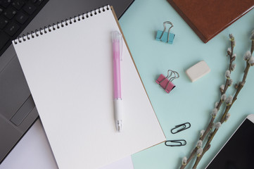 Concept for girl's desktop. On the desk is laptop, notepad for notes, phone,  pen, pencil, paper clips, eraser, office supplies, spring tree twigs. The background is turquoise with white. Flat lay.