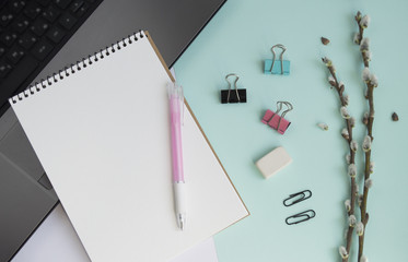 Concept for girl's desktop. On the desk is laptop, notepad for notes, phone,  pen, pencil, paper clips, eraser, office supplies, spring tree twigs. The background is turquoise with white. Flat lay.
