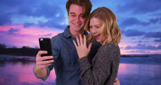 Happy engaged couple taking picture with cell phone on the beach at sunset
