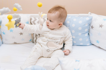 Cute baby sitting in a white round bed. Light nursery for young children.  Toys for infant cot. Smiling child playing with mobile of felt in sunny bedroom.