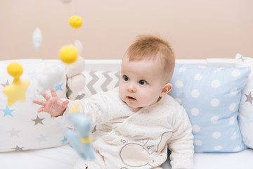 Cute baby sitting in a white round bed. Light nursery for young children.  Toys for infant cot. Smiling child playing with mobile of felt in sunny bedroom.