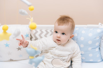 Cute baby sitting in a white round bed. Light nursery for young children.  Toys for infant cot. Smiling child playing with mobile of felt in sunny bedroom.