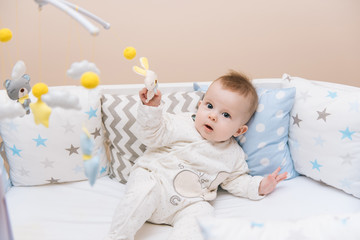 Cute baby sitting in a white round bed. Light nursery for young children.  Toys for infant cot. Smiling child playing with mobile of felt in sunny bedroom.