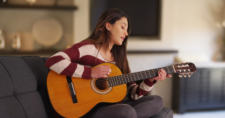 Portrait of cute Latina female lounging at home playing guitar and singing