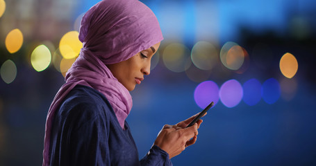 Black female in hijab texting on phone turning to smile at camera in evening