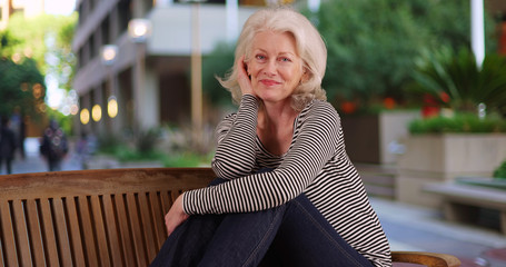 Portrait of mature Caucasian lady posing cheerfully on bench in outdoor courtyard