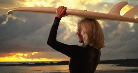 Mature white female surfer holding surfboard over head watching sunset at beach
