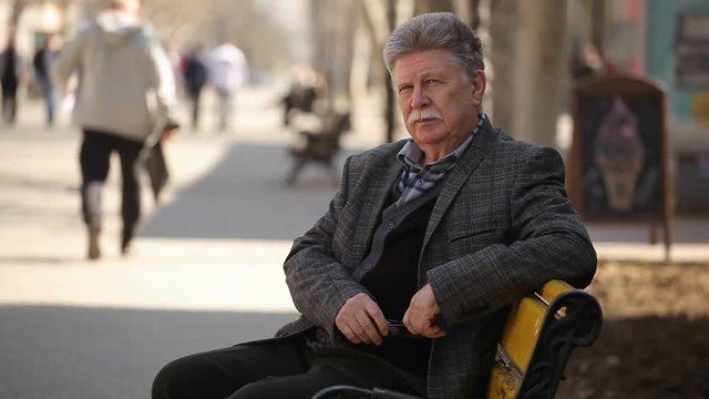 Profile of a confident senior man with a white mustache sitting on a bench and waiting for his wife in an alley on a sunny day in spring