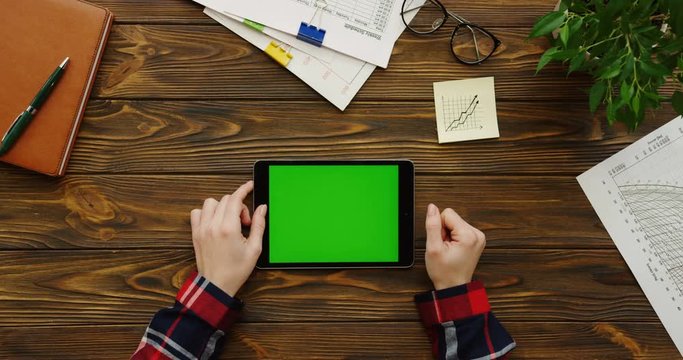 Close up of the female hands with the red plaid sleeves using the black tablet device with a green screen horizontally on the wooden office table with office stuff and plant. Chroma key. Top view