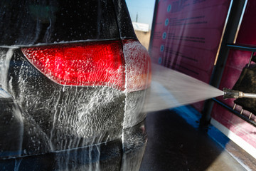 Naklejka premium Jet of wash with soap water high pressure is sent to the rear lights of the machine. Driver, girl wahsing her car in the stall of a car wash, using a high pressure water jet.