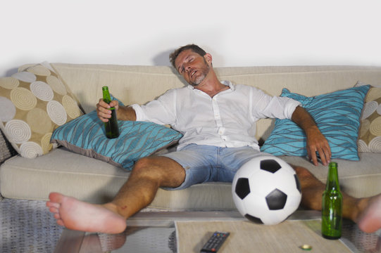 Young Wasted Soccer Supporter Man Lying On Home Sofa Couch Sleeping Drunk After Drinking Too Much Beer And Alcohol While Watching Football Match On TV