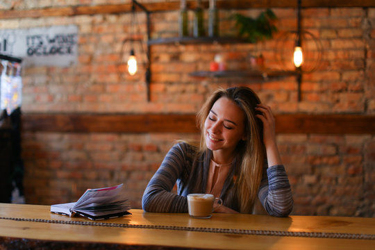 Young woman decided to devote time to herself on weekend and went to coffee house, smiling fair-haired female wearing white blouse and grey cloak sitting near wooden table with glass cup of drink and