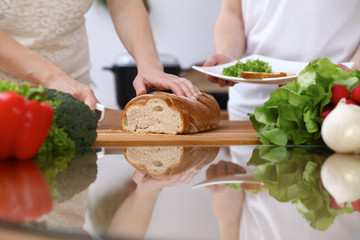 Obraz na płótnie Canvas Closeup of human hands cooking in kitchen. Mother and daughter or two female cutting bread at the table full of vegetables and green salad. Healthy meal, vegetarian food and lifestyle concepts