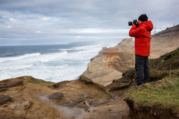 Obraz premium Adventurous man with a camera is standing taking pictures of the beautiful ocean during a vibrant winter morning. Taken in Cape Kiwanda, Pacific City, Oregon Coast, America.