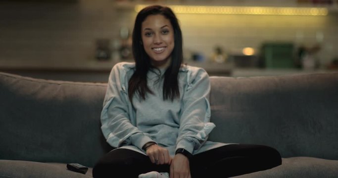 Girl sits down on a sofa in a cozy living room at night. She turns on the tv and smiles as her favourite movie starts.