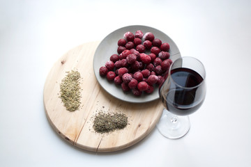 Glass of red wine, frozen cherries on grey concrete plate, rosemary and thyme spices on wooden cutting board isolated on white background