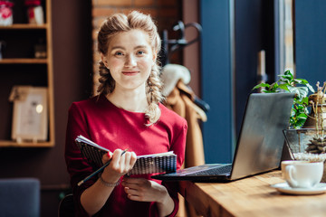 Portrait of Caucasian woman writting plans in notebook while sitting with laptop in modern loft cafe near window, female student freelancer thinking about new ideas during work on laptop computer.