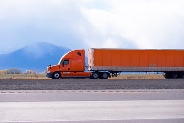 Orange big rig semi truck with long semi trailer running on the flat road with mountain on...