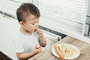 The child is sitting in the kitchen and greedily eats sausage