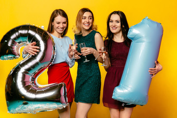 Young group of laughing girls in colorful dresses having fun together during celebrating birthday with helium balloons, holding a wineglasses with champagne, isolated on yellow.