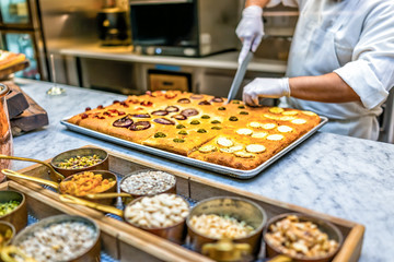 Man chef in restaurant kitchen cutting foccacia bread with knife, nuts, spices in containers and toppings after baking in oven, marble countertop