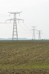 View of a row of steel lattice transmission towers supporting a high-voltage overhead power line across the french countryside, passing over plowed land under a pale light.