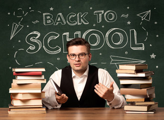 A young teacher in glasses sitting at classroom desk with pile of books in front of blackboard saying back to school drawing concept.