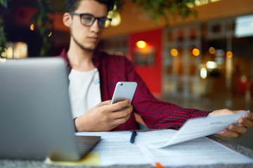 Fototapeta premium Smiling young hipster man in glasses with smartphone and laptop on the table. Programmer, web developer, designer working at office comparing mobile and desktop website versions. 