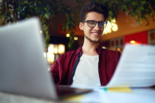 Smiling young hipster man in glasses with laptop on the table. Programmer, web developer, designer working at office comparing mobile and desktop website versions.
