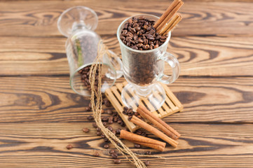 Lot of fried coffee beans in transparent glass for mulled wine on pallet near threads and three cinnamon sticks and coffee beans poured out of glass on old wooden rustic brown table