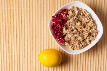 Cooked yummy warm oatmeal and heap of ripe fresh pomegranate seeds in white ceramic bowl near piece of pomegranate and whole lemon on clean bamboo rug with copy space