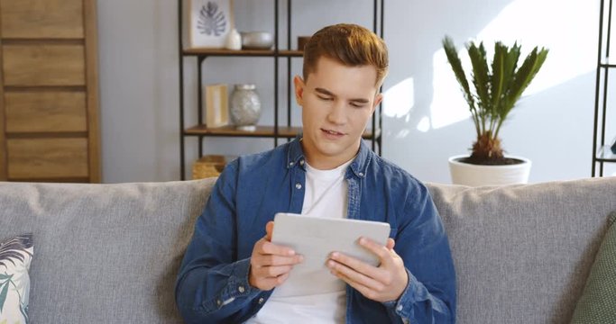 Close up of the attractive young man in the casual style scrolling on the tablet computer and smiling while sitting on the couch at home. Indoor