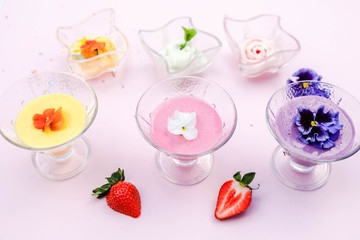 Ice cream in wave glass bowls and creamy milk beverage, smoothie with edible flowers *pansy, violet (on pink background. Pink (strawberry), yellow (mango or banana)  Top view