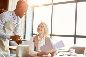Happy young businesswoman with menu looking at waiter while making order in cafe