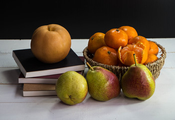 The Chinese Pear on books stacked and group of orange in woven basket put at the back of blurred green pears ,on timber board