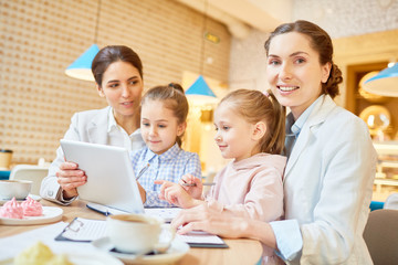 One of businesswomen looking at camera while sitting by table in cafe with her colleague and two little girls