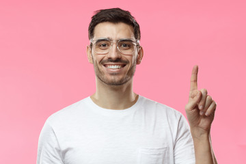 Attractive young man wearing transparent eyeglasses and white t-shirt pointing up with his finger isolated on pink background