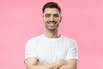 Obraz premium Portrait of smiling handsome man in white t-shirt and transparent eyeglasses standing with crossed arms isolated on pink background