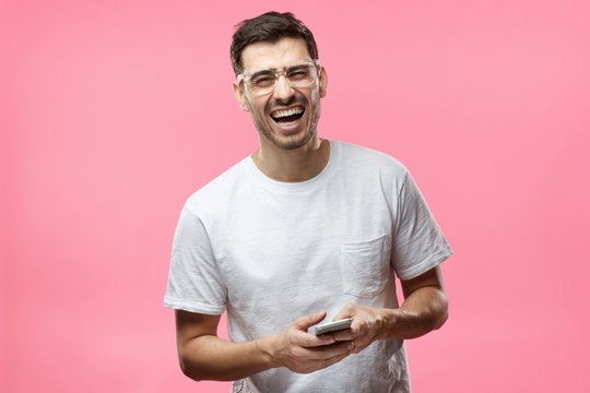 Portrait of handsome young man in white t-shirt, holding smartphone, looking at camera and laughing, isolated on pink background