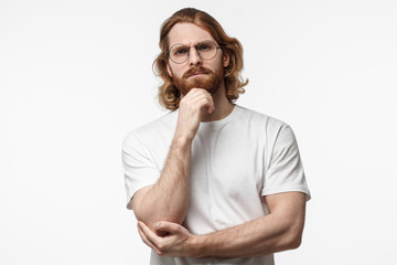 Skeptical and dissatisfied young brural bearded man in white tshirt touch his chin with hand. Doubt concept