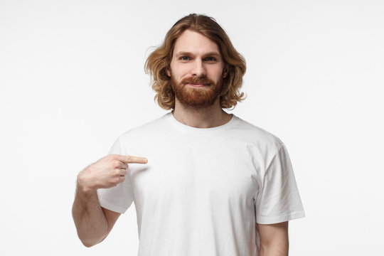 Young bearded man pointing with index finger at blank white tshirt with empty space for your advertising text or image, standing isolated on gray background