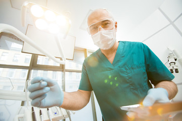 Contemporary dentist in protective gloves, mask, eyeglasses and uniform going to examine oral cavity of patient with instrument