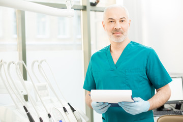 Aged dentist in uniform and gloves looking at camera while standing by his workplace with dentistry equipment