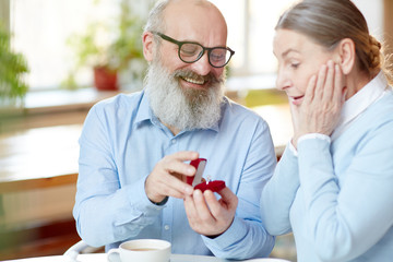 Astonished senior woman looking at expensive ring in small velvet box held by her husband