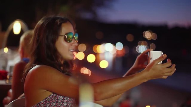 Pretty brunette woman in sunglasses posing making faces taking selfie photo using smartphone with flash and bokeh lights in beach cafe. 1920x1080