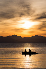 Naklejka na ściany i meble Silhouettes of two men rowing in a small fishing boat at a colorful golden cloudy sunrise in Corfu, Greece. Vertical image