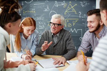 Confident teacher explaining details of his lecture points to group of students after lesson