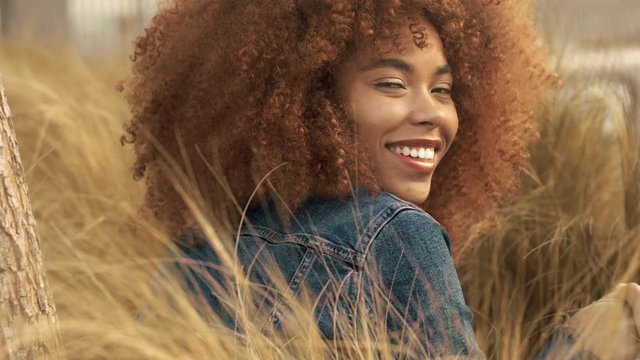 black mixed race woman with big afro curly hair sits on lawn with high grass on sunset. Denim look on hay