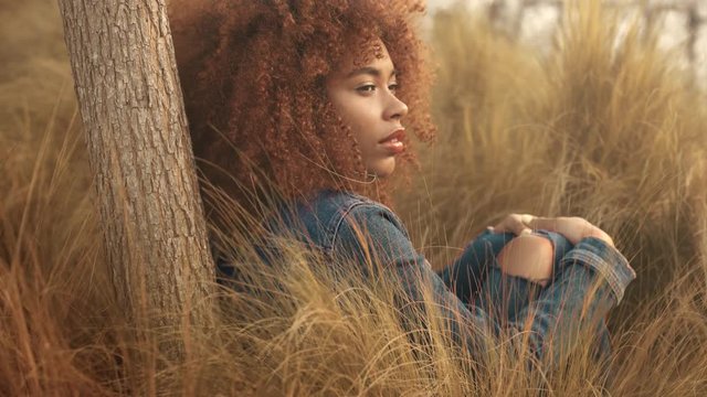 black mixed race woman with big afro curly hair sits on lawn with high grass on sunset. Denim look on hay