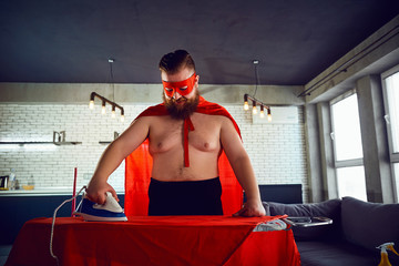 Thick funny man in a superhero costume ironing things at home. Household chores.